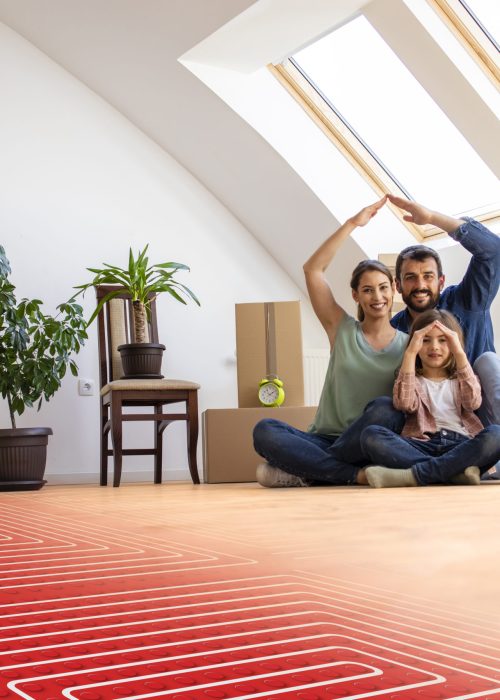 Home, people, moving and floor heating concept - portrait of happy family sitting on warm parquet with floor heating and pipes.